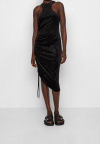 Modelka stojí v úzkých, černých šatech, které přiléhají k tělu a mají strukturovaný vzor a vysoký límec, doplněná o robustní černé sandály.