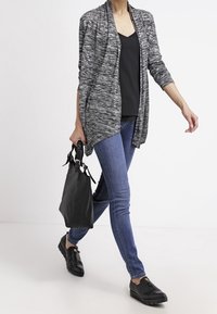 Femme marchant en jeans bleus, top noir, cardigan ouvert gris, chaussures noires, et portant un sac à main noir dans la main droite.