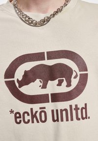 Bežinė medvilninė marškinė su tamsiai rudos spalvos rinokero grafika, esančia apvalaus stačiakampio logotipe, derinama su storu sidabriniu grandinės kaklo papuošalu.