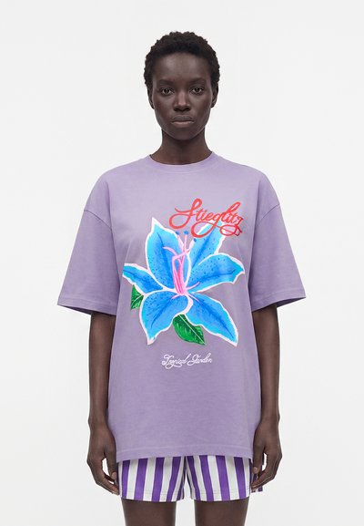 Femme portant un T-shirt lavande oversize avec une grande fleur bleue et un texte rouge, associé à un short rayé violet et blanc, debout devant un fond blanc.