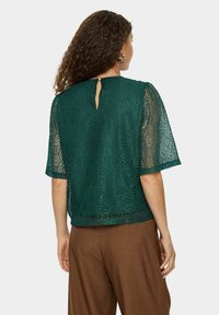 Haut en dentelle teal à manches courtes, avec un dos en trou de serrure et un motif texturé. Associé à un pantalon marron, mettant en valeur les couleurs contrastées.