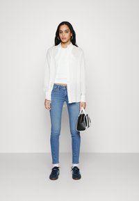 Vit skjorta med knappar, åtsittande vit linne, blå skinny jeans och marinblå sneakers. Håller en liten svart och vit väska.