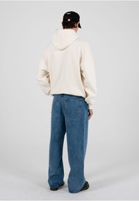 Persoană care poartă un hanorac crem, blugi largi albastre, pantofi negri și o șapcă neagră, stând pe un fundal simplu și deschis la culoare.