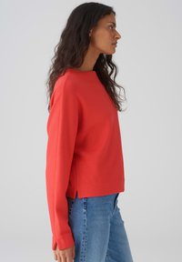 Sweatshirt rouge avec un col rond, des manches longues, des fentes latérales et une coupe décontractée, associé à un jean en denim bleu.