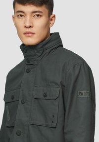 Grau geblümte Jacke mit hohem Kragen, Knopfverschluss vorne und zwei Brusttaschen mit Klappe und Knopfdetail. Strukturiertes Material mit einem Etikett am Ärmel.