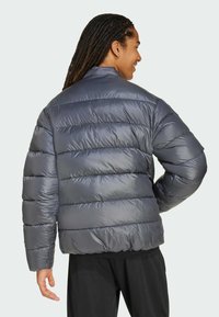 Osoba z długimi, ciemnymi włosami, ubrana w ciemną, puchową kurtkę, widziana od tyłu na jasnoszarym tle.