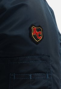 Tissu bleu marine avec une poche cousue et un petit patch noir en forme de bouclier arborant les lettres rouges "KC" bordées de fil jaune.