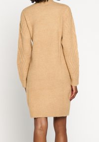 Robe pull beige tricotée avec un col montant, des manches longues à torsades et une taille cintrée, terminée au-dessus du genou par un ourlet côtelé.
