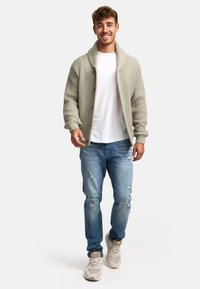 Cardigan gris de punto con cuello chal, camiseta blanca, jeans claros desgastados y zapatillas beige, mostrado en un modelo masculino sonriente.