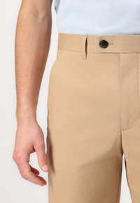 Pantaloni bej dintr-un material ușor, cu un design clasic, se închid cu un buton în față și au buzunare laterale, având o textură netedă.