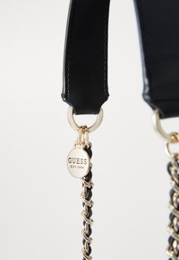Cinturino in pelle nera con hardware dorato, dotato di un'etichetta circolare con la scritta "GUESS EST. 1981." Il dettaglio a catena aggiunge texture e interesse visivo.
