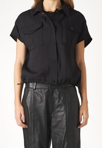 Chemise noire à manches courtes avec deux poches frontales, un col boutonné et un ourlet élastique froncé. Fabriquée en tissu léger, offrant une coupe décontractée.