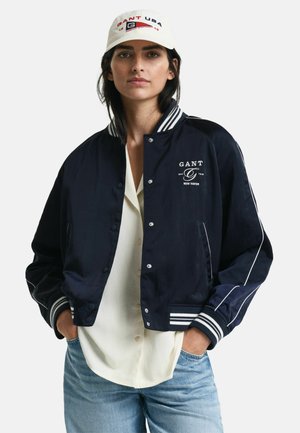 Navy bomber jacket with white stripes on the collar and cuffs, snap buttons, and GANT logo. Worn over a white shirt and paired with jeans.