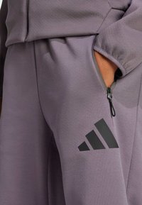 Pantalones deportivos morados hechos de un material suave, con un bolsillo con cremallera y un logo negro de Adidas cerca del bolsillo.