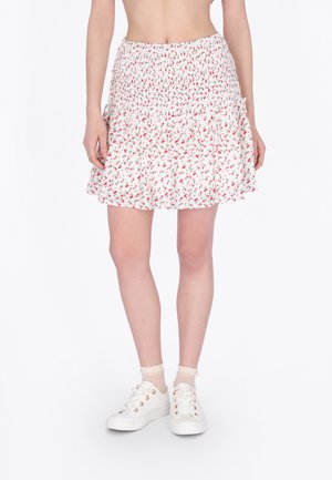 Frau trägt einen weißen geblümten gerafften Minirock kombiniert mit transparenten Knöchelsocken und weißen Turnschuhen, steht vor einem weißen Hintergrund.