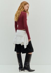 Bordeauxrotes Langarmshirt, weißer, schichtweise um die Hüften gewickelter Stoff, schwarze kniehohe Stiefel und schwarze knielange Shorts. Glatte Texturen, schlichtes Design.