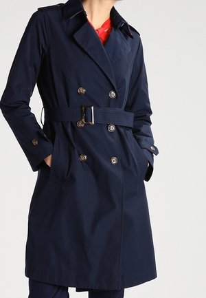 Kvinde iført en marineblå dobbeltradet trenchcoat med bælte i taljen og hænder i lommerne over en rød top.