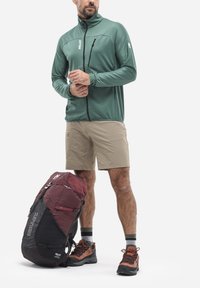 Grüne zip-up Jacke mit strukturiertem Finish, beiger Shorts und braunen Wanderschuhen. Roter und schwarzer Rucksack mit verstellbaren Trägern.