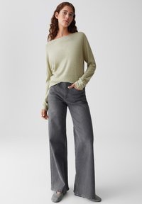 Femme avec de longs cheveux bouclés portant un haut à manches longues texturé vert clair, un jean gris taille haute à jambes larges et des chaussures gris-ardoise à enfiler.