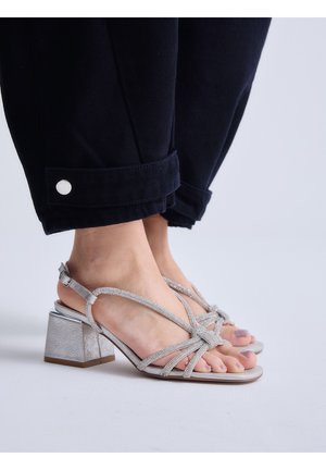 Pies con sandalias plateadas de tacón grueso y tiras, combinadas con pantalones negros con puños abotonados sobre un fondo claro.