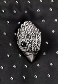 Spilla argentata a forma di uccello con piume lavorate e accenti di pietre nere, posata su un tessuto nero punteggiato da perline argentate.