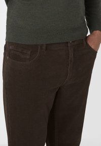 Person in dunkelbraunen Cordhosen, mit Händen in den Taschen, und einem olivgrünen Pullover, sichtbar bis zur Taille.