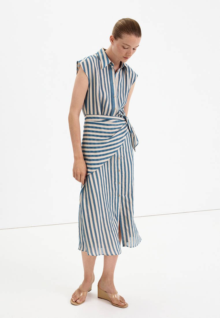 Femme portant une robe midi sans manches à rayures verticales bleues et blanches avec des boutons et une taille nouée, debout et regardant vers le bas.