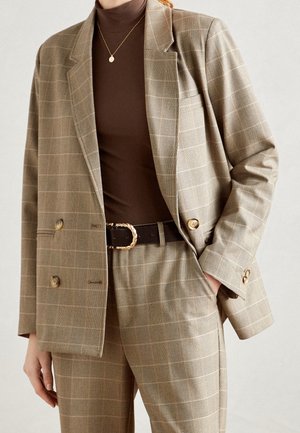 Person wearing beige plaid suit with double-breasted jacket, high-waisted matching pants, dark brown turtleneck, black belt with gold buckle, and gold necklace.