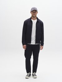 Marinblå jacka med dragkedja över en vit t-shirt, matchande marinblå byxor och en grå keps med en logotyp. Vita sneakers med svarta detaljer. Minimalistisk design.