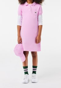 Růžové polo šaty s krátkými rukávy, n�áprsní kapsou a odpovídající růžovou čepicí. Zeleno-růžové pruhované ponožky a bílé tenisky dotvářejí vzhled.