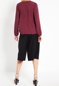 Blouse bordeaux à manches longues avec un col rond, associée à un pantalon culotte noir, portée avec des sandales blanches à talons. Finition en tissu lisse.