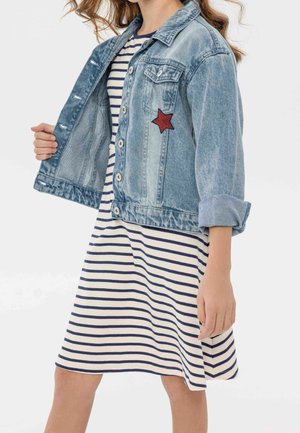 Džínová bunda světle modré barvy s ohrnutými rukávy a červenou hvězdnou nášivkou, nošená přes proužkované námořnické a bílé šaty. Tlačítka viditelná na přední straně.