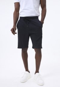 Pantalones cortos negros texturizados con una cintura elástica y cordón, combinados con zapatillas blancas. Los pantalones cortos tienen bolsillos laterales y un ajuste relajado.