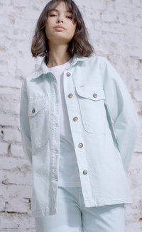 Jaqueta de ganga verde menta clara com dois bolsos frontais, fechos de botão e um corte descontraído, combinada com uma camisa branca e calças.