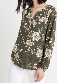 Blomsterbluse med V-udskæring, lange ærmer og en afslappet pasform. Mørkegrønt stof prydet med hvide og gule blomster.