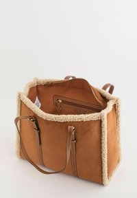 Sac fourre-tout en daim beige avec bordure en polaire crème, deux poignées en cuir brun, et une fermeture éclair. L'intérieur comprend une poche supplémentaire zippée.