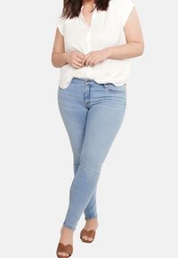 Woman wearing light blue skinny jeans, white short-sleeve blouse, and brown open-toe sandals standing with legs crossed.