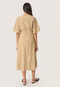 Beige klänning med korta, utsvängda ärmar, prydd med utskärningar och en midilång kjol. Slät textur och dragkedjestängning bak.