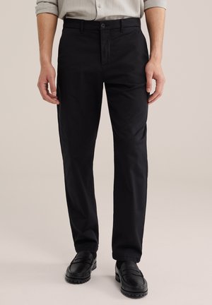 Mees, kes kannab kitsaid musti pükse, musti loafereid ja helekat värvi üleskeeratud varrukatega särki, seisab ühevärvilise tausta ees.