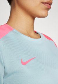Top deportivo de Nike con un cuerpo azul claro, acentos en rosa en los hombros y un logo Swoosh en rosa. Textura suave con detalles de patrón acanalado.