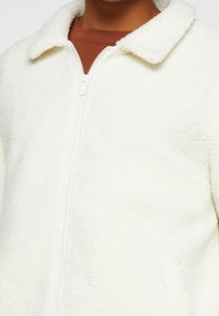 White fleece jacket with a zip closure, featuring a classic collar and textured, soft material. Shows a brown inner layer beneath.