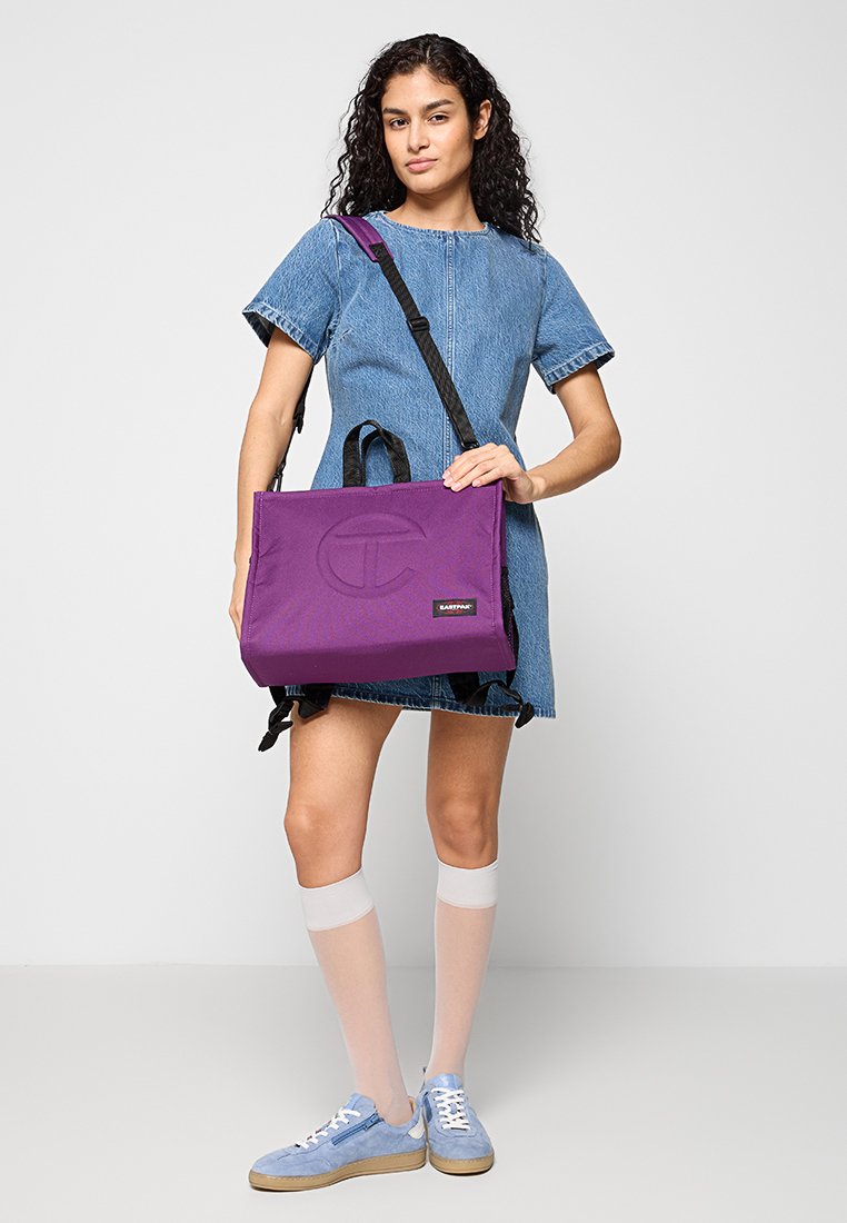 Borsa a tracolla in tessuto viola con logo testurizzato, tracolla regolabile e doppi manici. Il modello indossa un vestito di denim e scarpe sportive.
