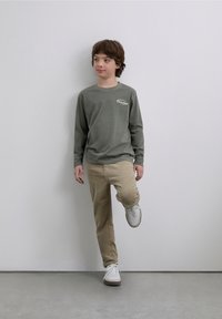 Grå T-shirt med långa ärmar och en logga på bröstet, kombinerad med khakifärgade byxor och vita sneakers. Bakgrunden är enkel och neutral.