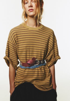 Randig gul t-shirt med uppvikta ärmar, med vinröda ränder. Bärs över ett blått bälte och en svart underdel. Avslappnad, lös passform.