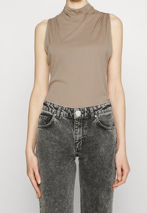 Kobieta ubrana w bezrękawnik o kolorze taupe, wpuszczony w wysokowysokie, czarne jeansy z efektem kwasowego prania, z przednimi kieszeniami i metalowym zamknięciem na guzik.