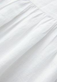 Tissu en coton blanc avec une texture douce et lisse, présentant des plis froncés sur un bord, créant une dimension subtile. Apparence légère et aérée.
