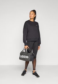 Μαύρο φούτερ με κεντημένο λογότυπο, σε συνδυασμό με μαύρο κολάν και αθλητικά παπούτσια. Μια μαύρη τσάντα duffle με λευκό λογότυπο "adidas" κρατιέται στο δεξί χέρι.