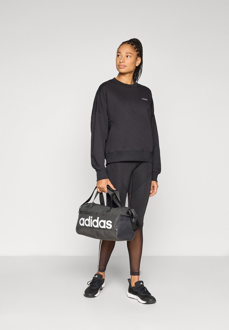 Sweat-shirt noir avec logo brodé, associé à des leggings noirs et des baskets. Un sac de sport noir avec logo "adidas" blanc est tenu dans la main droite.