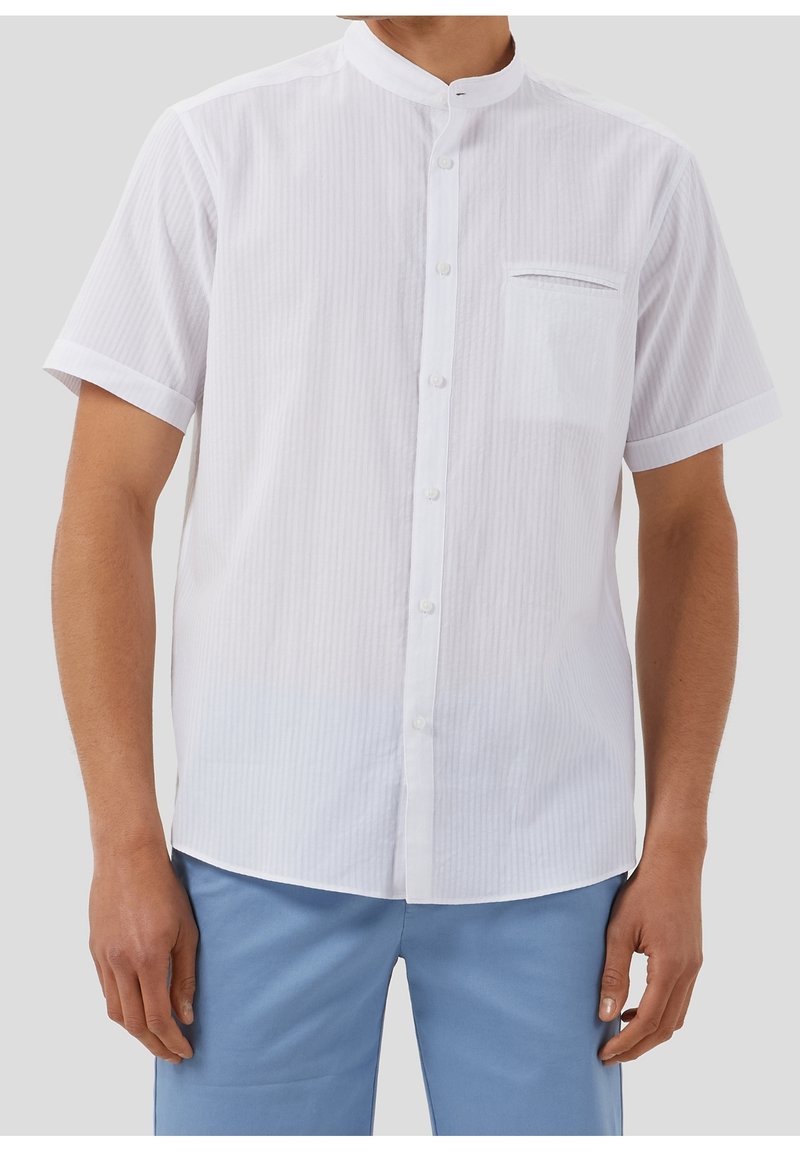 Uomo che indossa una camicia bianca a maniche corte con colletto alla cinese, bottoni frontali e taschino sul petto, abbinata a pantaloni azzurri chiari.