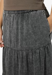 Pilkos džinsinio audinio sijonas su keliais sluoksniais, turintis elastinį juosmenį, vertikalių tekstūrų ir šonines kišenes. Minkšta, kasdieniška medžiaga su šiek tiek blizgesio.